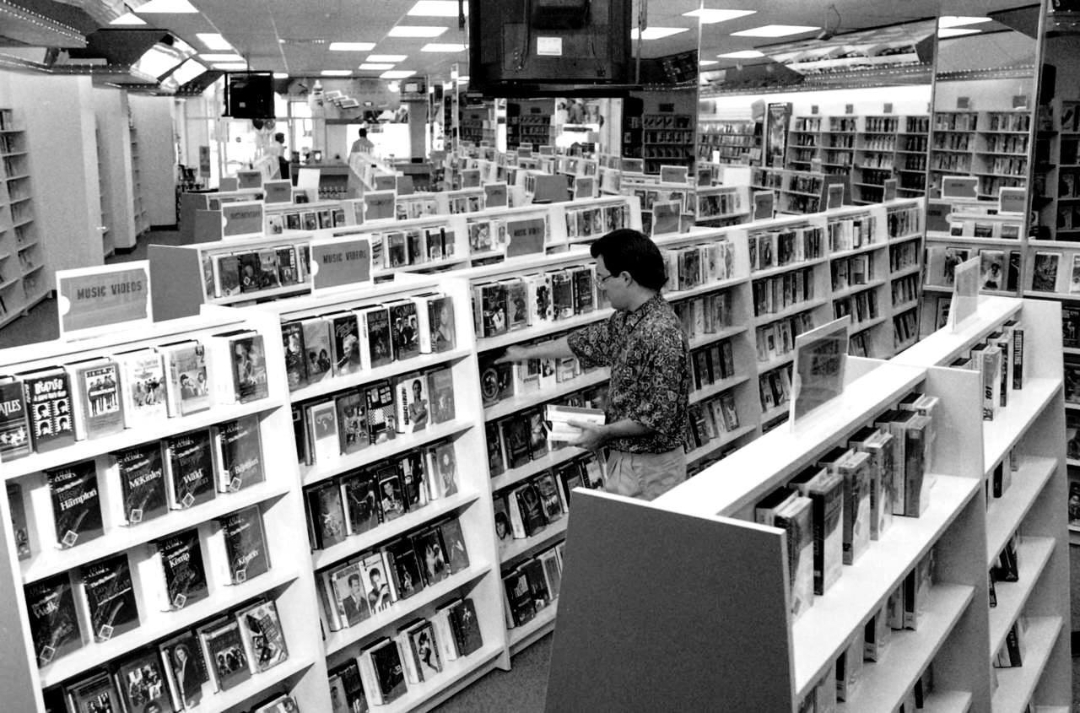 A person stands in an aisle browsing VHS tapes on shelves in a large, well-lit video rental store filled with rows of tapes and signs labeled “Music Videos.”