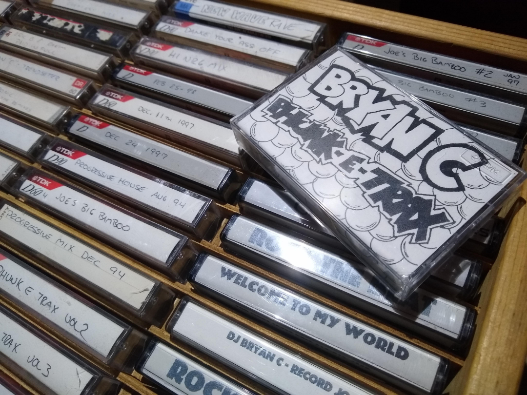 A close-up of a drawer filled with labeled cassette tapes. One tape with a hand-drawn label reading "BRYAN C HAMMER TRAX" is prominently displayed on top of the others.