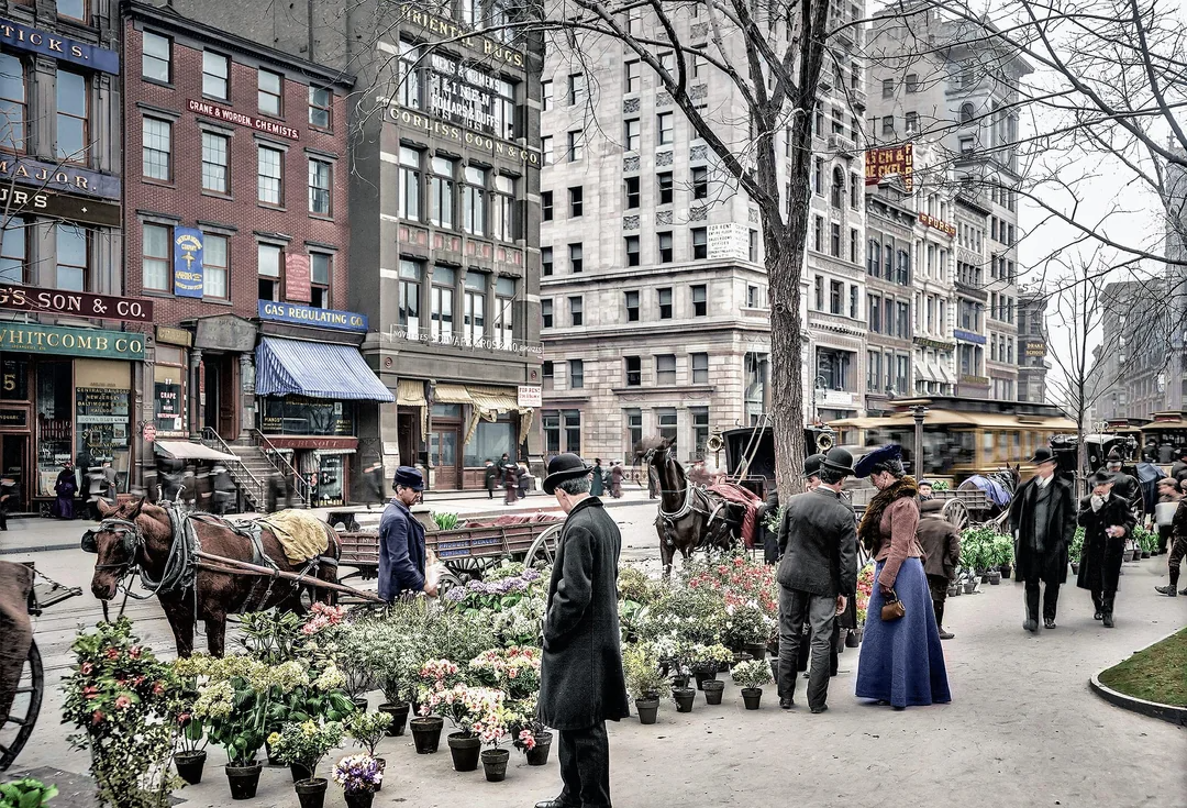 People shop for flowers at an outdoor market on a city street with horse-drawn carriages and early 20th-century buildings in the background. Trees line the sidewalk and people wear period clothing.