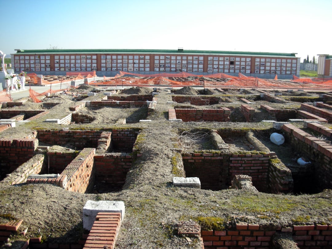 Excavated brick ruins at an archaeological site, with orange safety netting and a long wall of cemetery niches in the background under a clear sky.