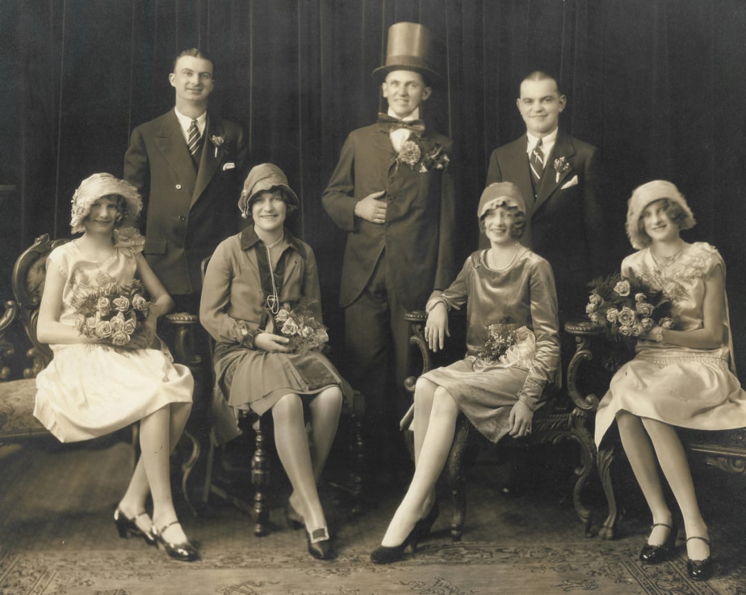A vintage black-and-white photo of four women in dresses and hats holding bouquets, seated on ornate chairs, with three men in suits standing behind them; one man wears a top hat. The group poses in front of a dark curtain.