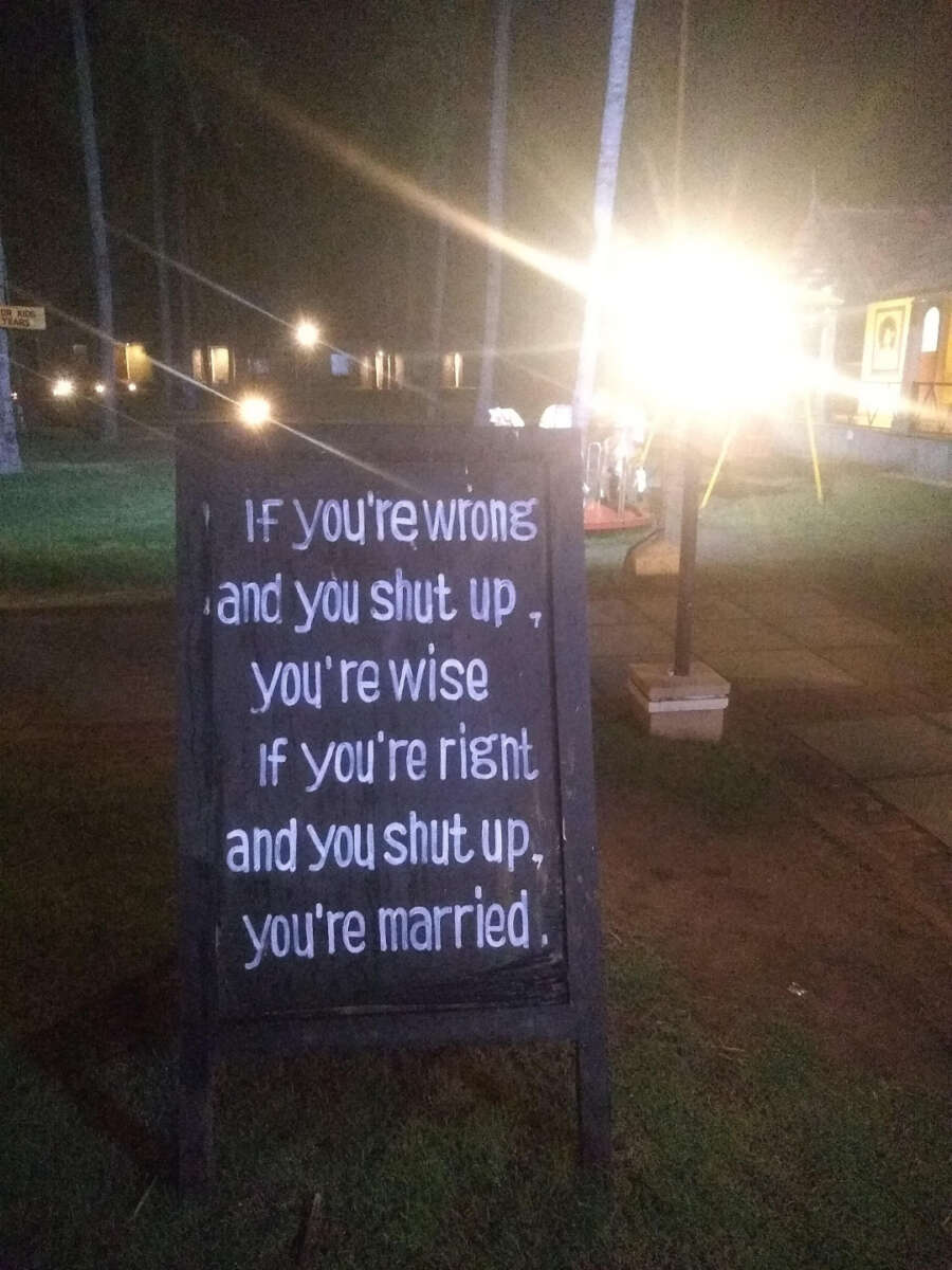 A chalkboard sign outdoors at night reads: "If you're wrong and you shut up, you're wise. If you're right and you shut up, you're married." Bright lights and palm trees are visible in the background.