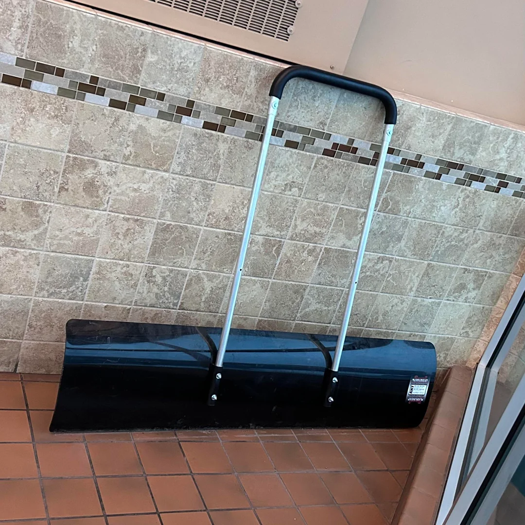 A large push-style snow shovel with a black blade and silver handle is leaning against a tiled wall next to a glass door.
