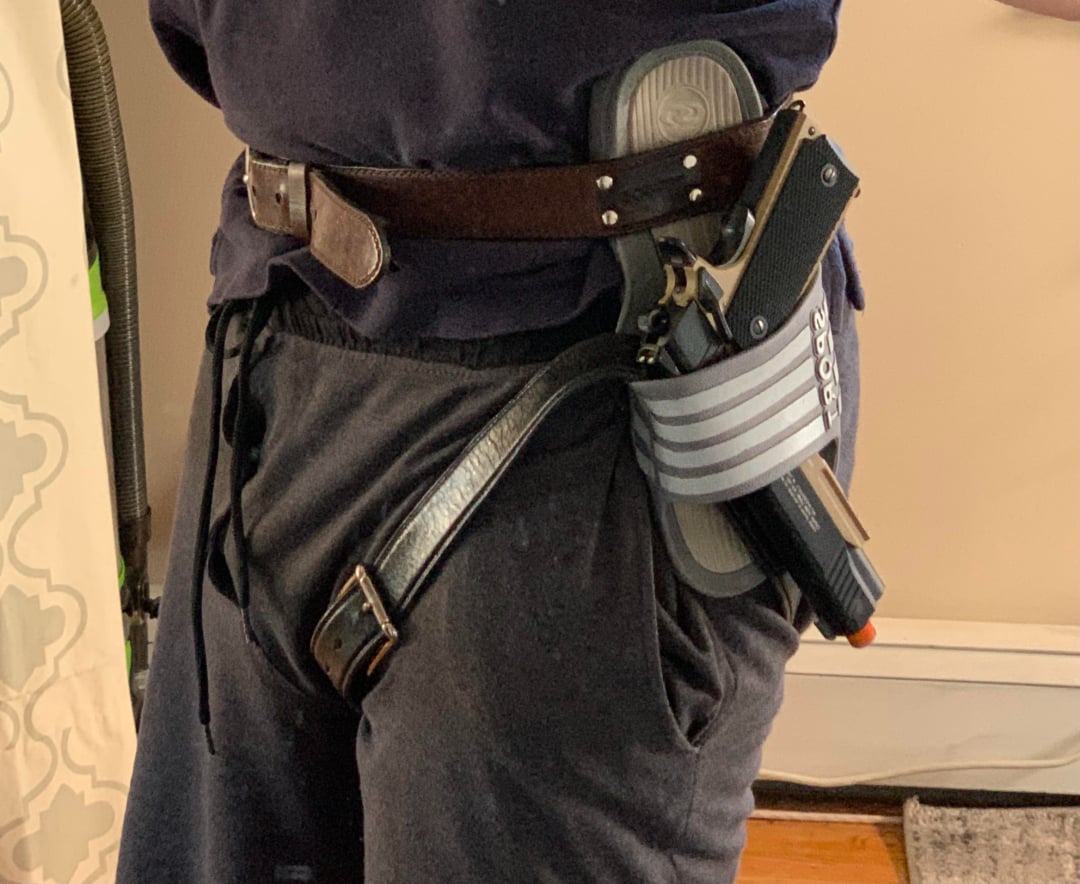 A person wearing a dark shirt and sweatpants has a handgun in a holster attached to their belt, with an extra belt loosely hanging down.