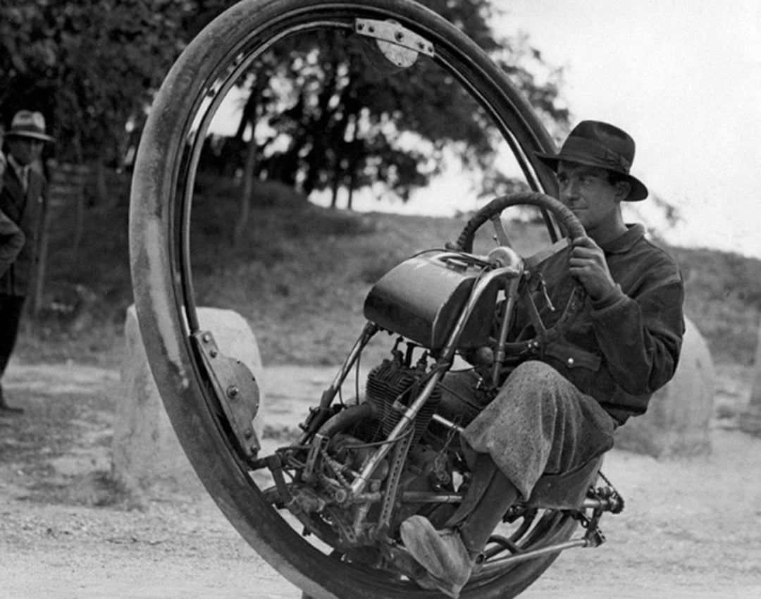 A man wearing a hat and coat rides a large monowheel motorcycle, sitting inside its single wheel, with trees and another person visible in the background.