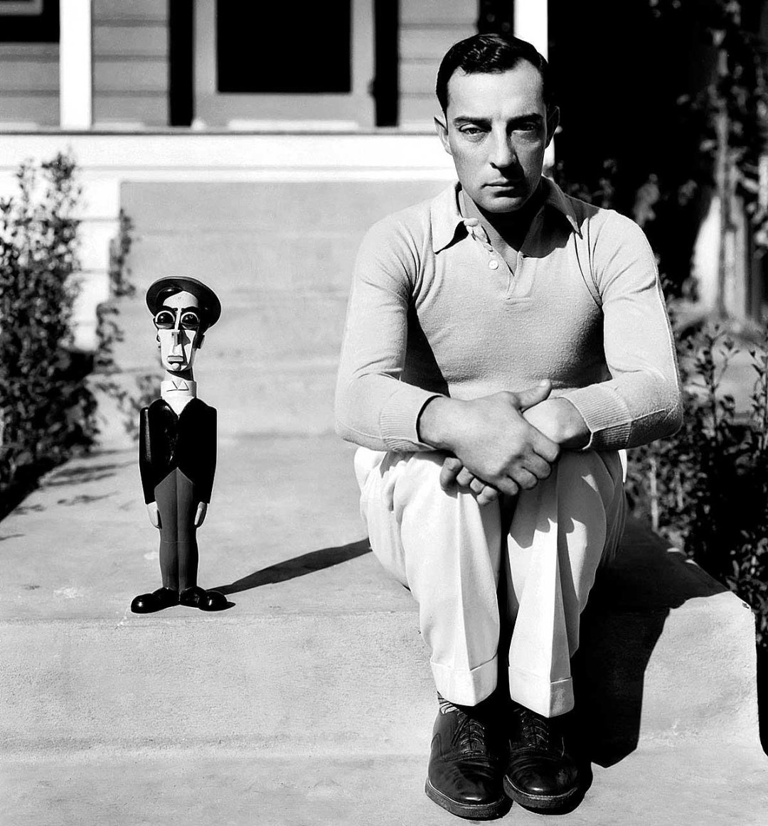 A man sits on outdoor steps with his arms resting on his knees, looking serious. Beside him is a wooden doll dressed in similar clothing. The background shows part of a house with a porch and stairs.