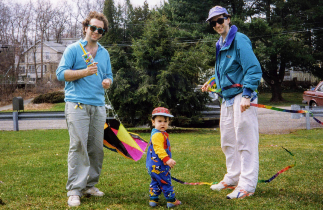 Two adults and a young child stand on grass, holding colorful kites. The adults wear sunglasses and casual clothes, while the child in a cap and overalls looks at the camera. Trees and houses are visible in the background.