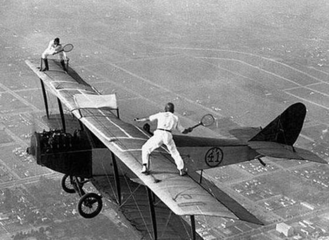 Two people in tennis attire play a game of tennis on top of a flying biplane, each standing on a wing with rackets, high above a city landscape.