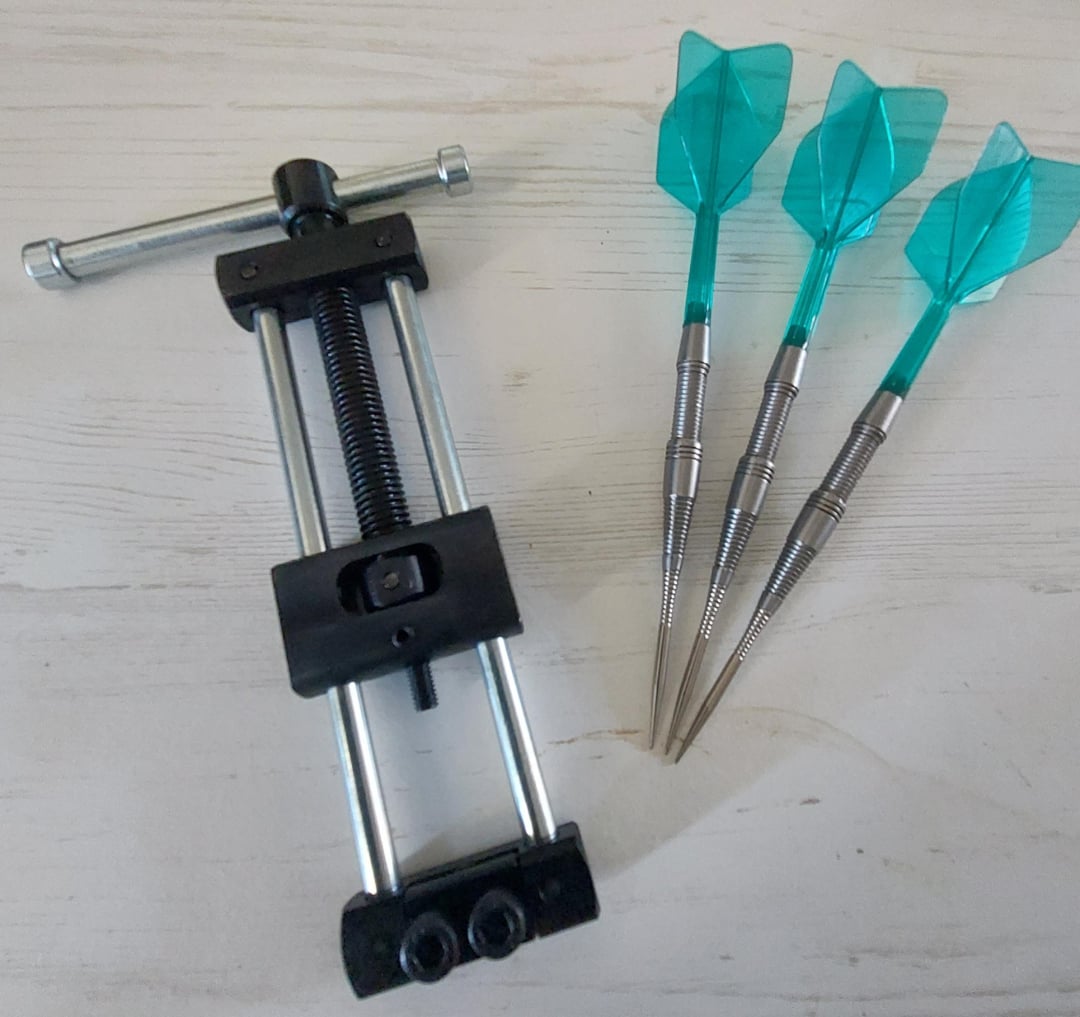 A metal tool with a screw mechanism lies next to three turquoise darts with metallic tips and flights, all placed on a light-colored wooden surface.