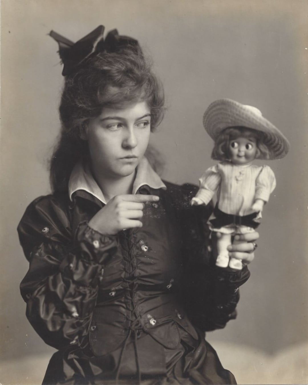 A young woman in vintage clothing and a large bow in her hair points at a doll she is holding up. The doll wears a dress and a wide-brimmed hat. The image is in black and white.