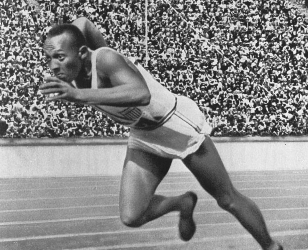A Black male athlete in a sleeveless white uniform starts a sprint on a track, with a packed stadium of spectators in the background. The image is black and white, capturing a moment of intense focus and action.