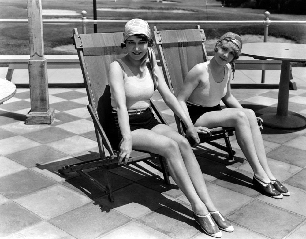 Two women in vintage swimsuits and swim caps sit on deck chairs on a tiled patio, holding hands and smiling. The scene appears to be from the early 20th century, with outdoor furniture and grass in the background.