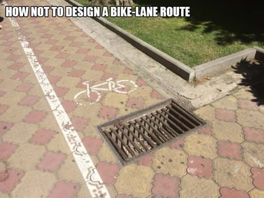 A bike lane painted on pavement leads directly into a large, unsafe storm drain grate. Text at the top reads, "HOW NOT TO DESIGN A BIKE-LANE ROUTE.