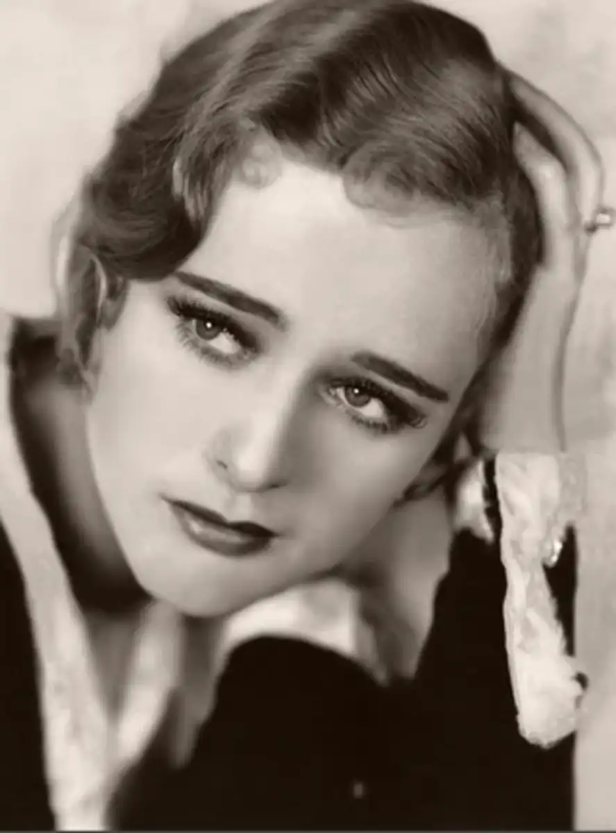 A black-and-white portrait of a woman with wavy hair, gazing upward with a pensive expression, resting her head on her hand. She wears dark lipstick and a collared garment.
