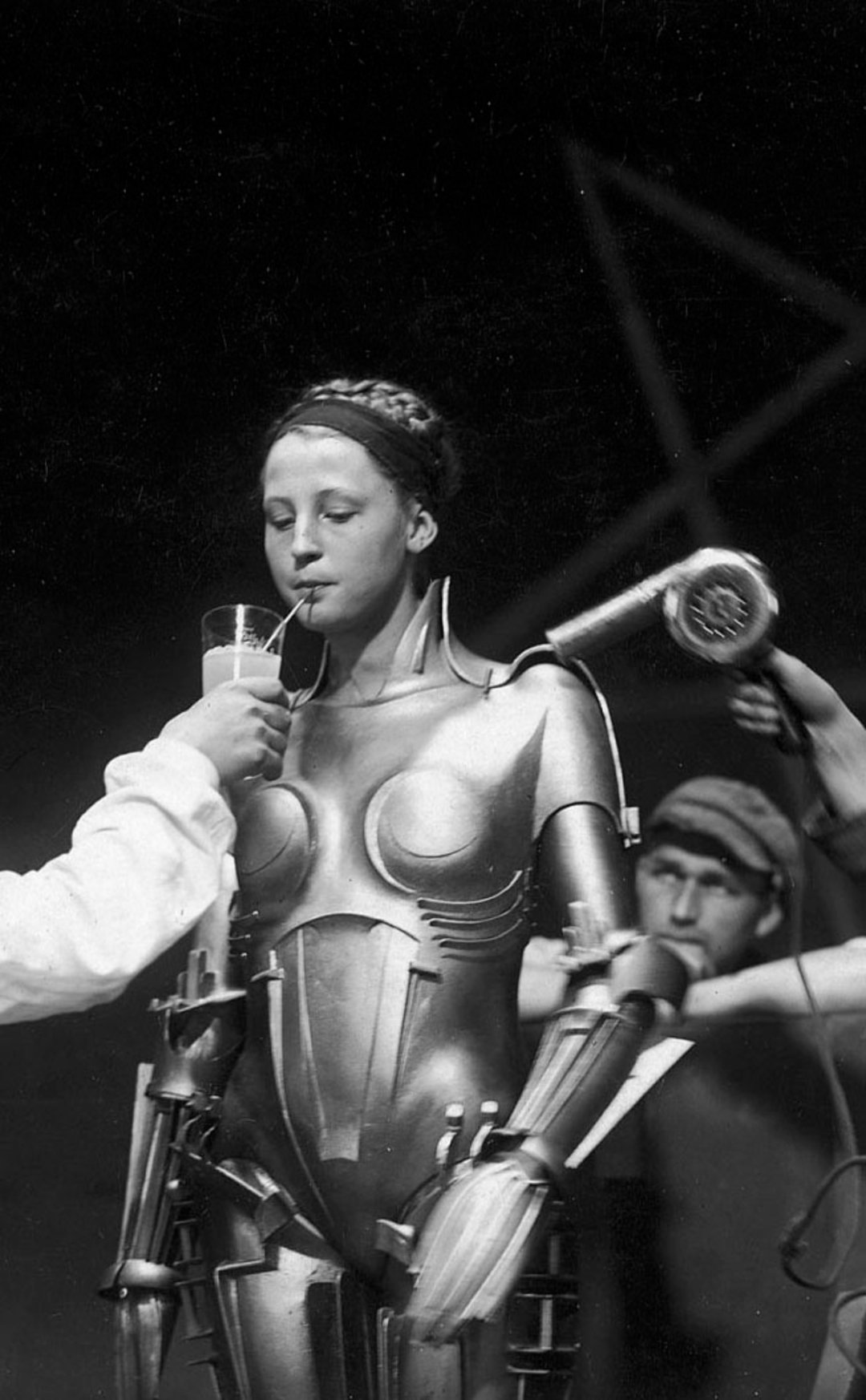 A woman in a metallic robot costume drinks from a glass with a straw while someone holds a hair dryer near her; another person adjusts her costume in the background.