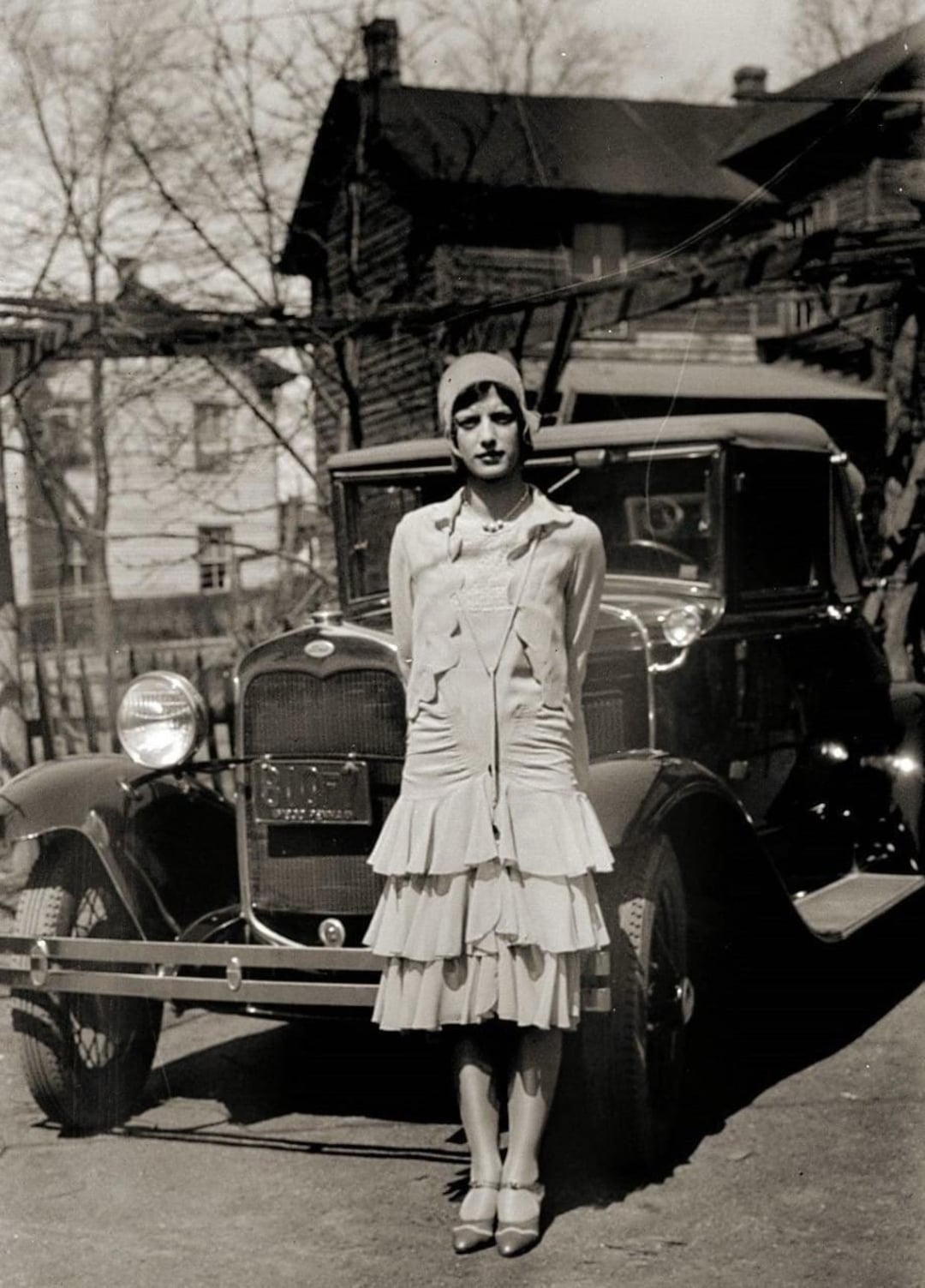 A woman in a 1920s flapper dress and cloche hat stands in front of a vintage car on a sunny day, with houses and bare trees in the background.