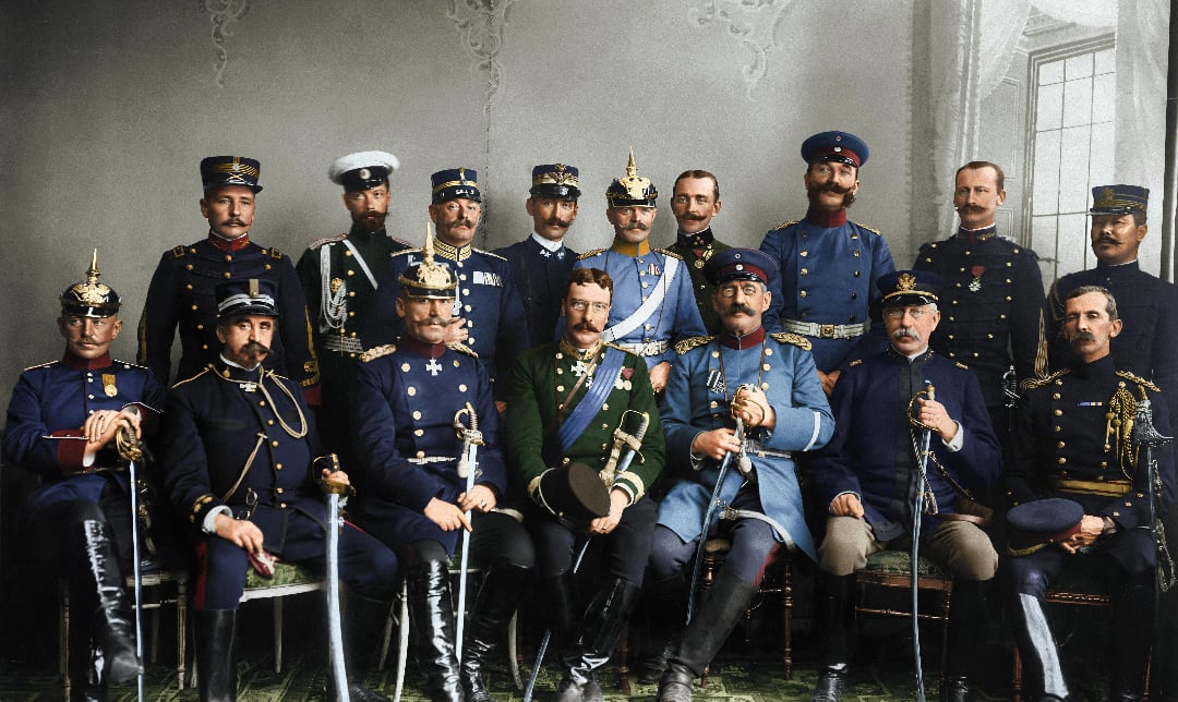 A group of men in elaborate military uniforms, some seated and others standing behind them, pose together in a formal indoor setting with ornate decor and large windows.