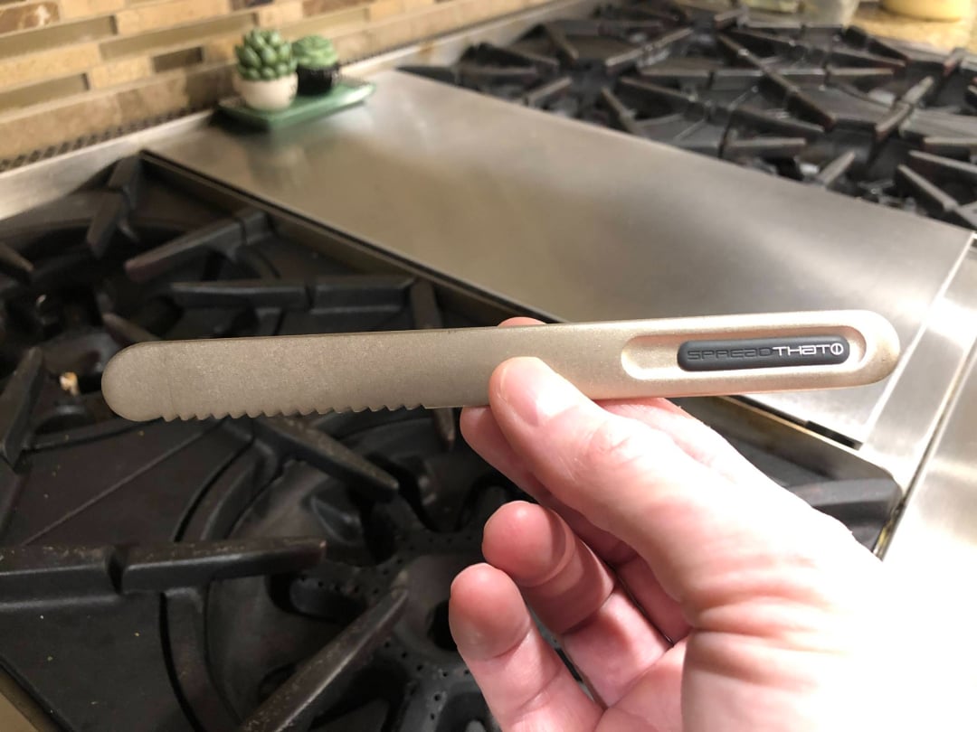 A hand holds a gray, serrated utensil labeled "SPREAD THAT! PRO" in a kitchen with a gas stovetop and small potted succulents on the counter in the background.