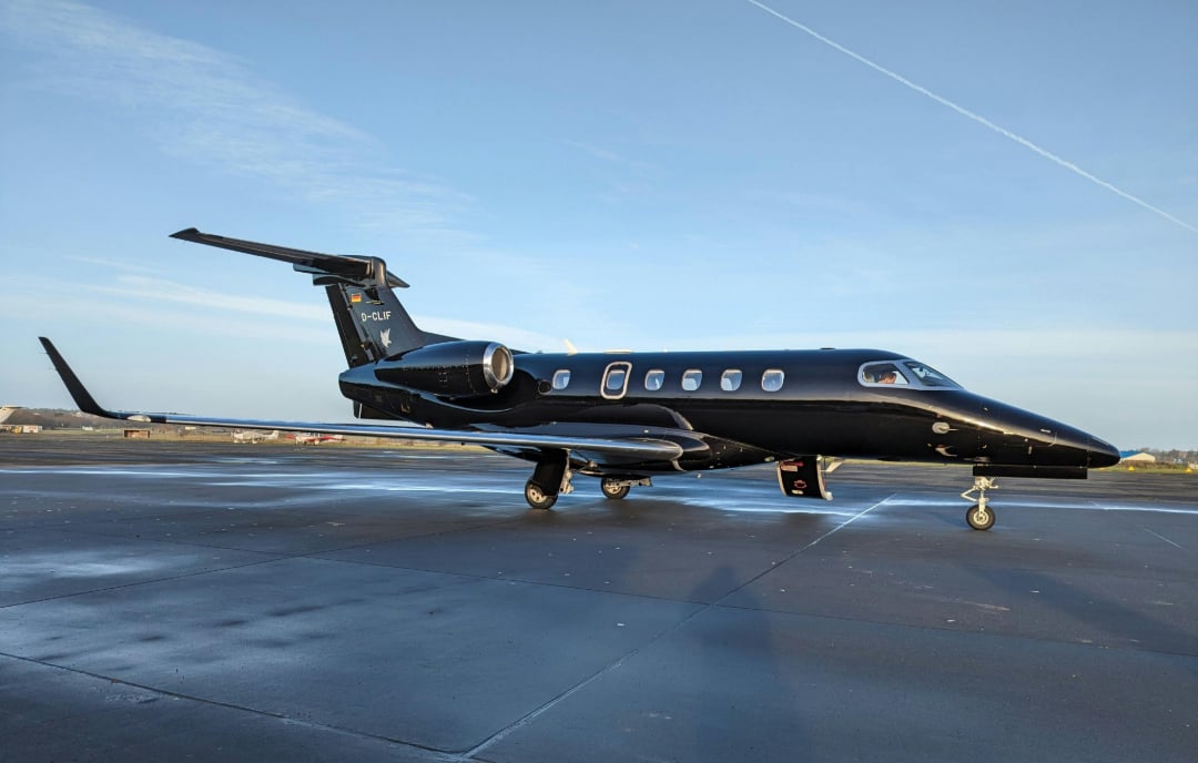 A sleek black private jet is parked on an airport tarmac under a clear blue sky, with its entry door open and runway markings visible on the ground.