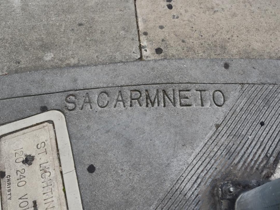 Concrete sidewalk with the word "SACARMNETO" misspelled and etched into the surface; nearby are utility markings and tactile paving for accessibility.