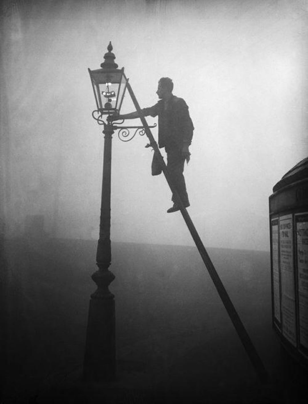 A person standing on a tall ladder lights a streetlamp in thick fog, creating a silhouette effect against the misty background.