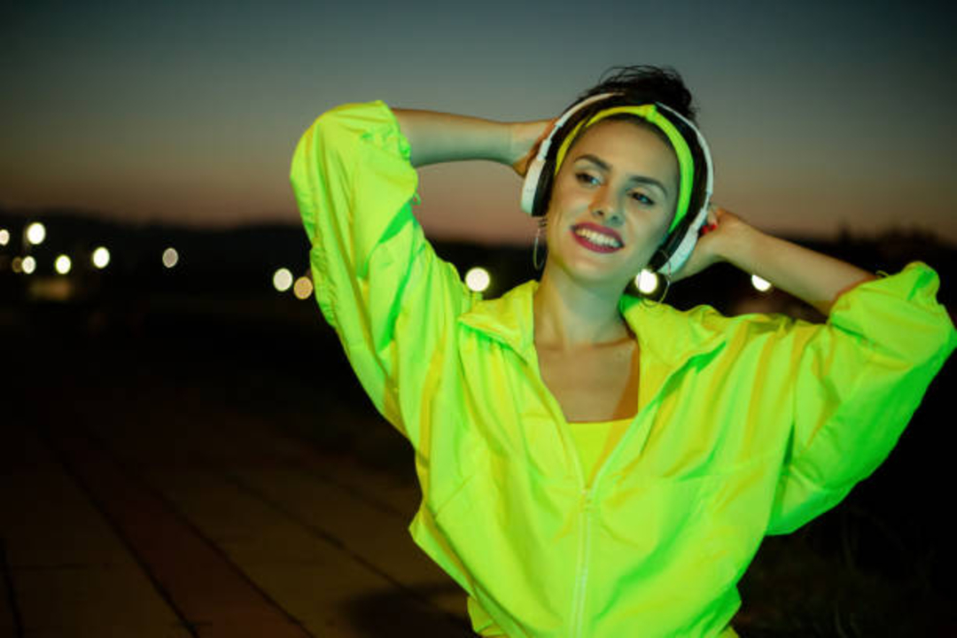 A smiling woman wearing a bright neon yellow jacket and headband listens to headphones outdoors at dusk, with blurry city lights in the background.