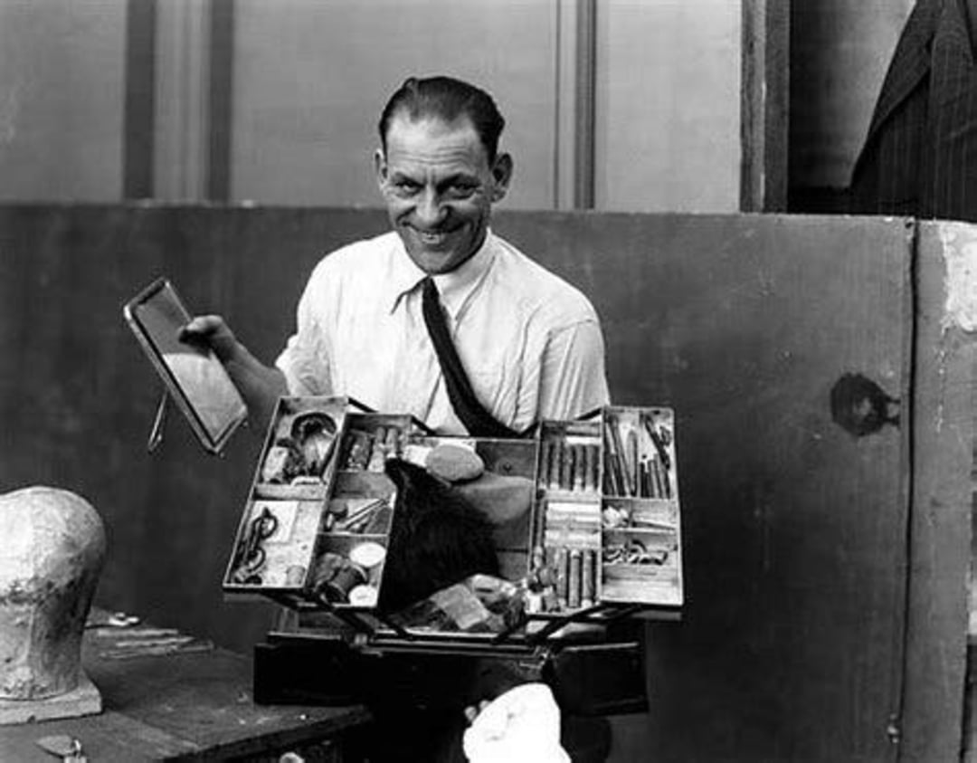 A man in a white shirt and loosened tie smiles while holding an open makeup kit filled with brushes and cosmetics; a mannequin head is visible in the foreground.