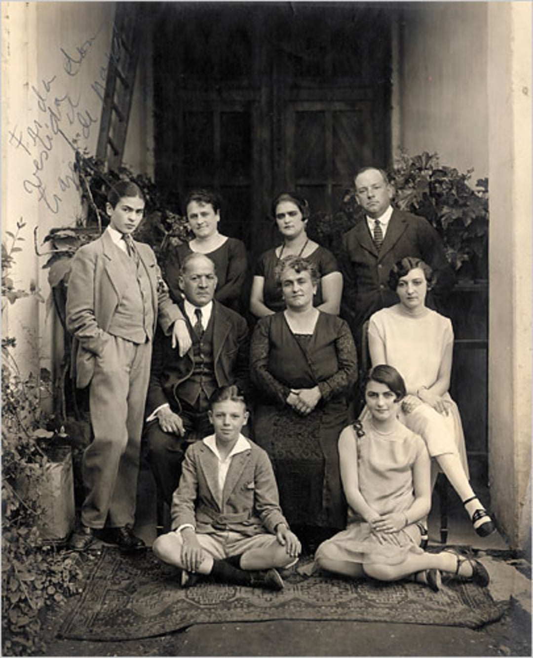 A vintage black-and-white photo of nine people, including men, women, and children, posed in front of a building entrance; some are seated and some are standing, dressed in formal early 20th-century clothing.