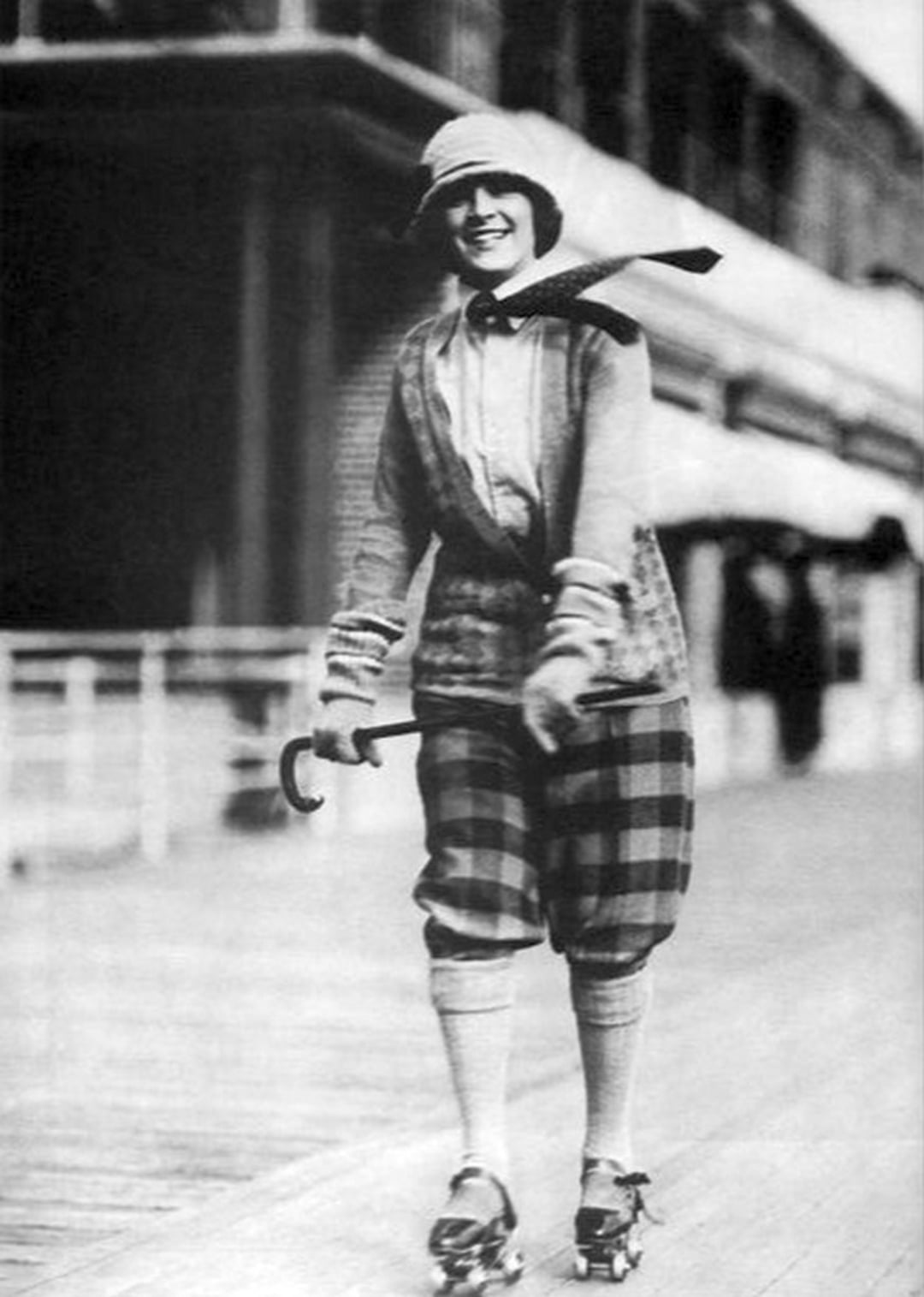 A woman wearing a cloche hat, scarf, gloves, and plaid knickerbockers roller skates on a boardwalk, smiling and holding a small cane. The background is blurred, showing buildings and railings.