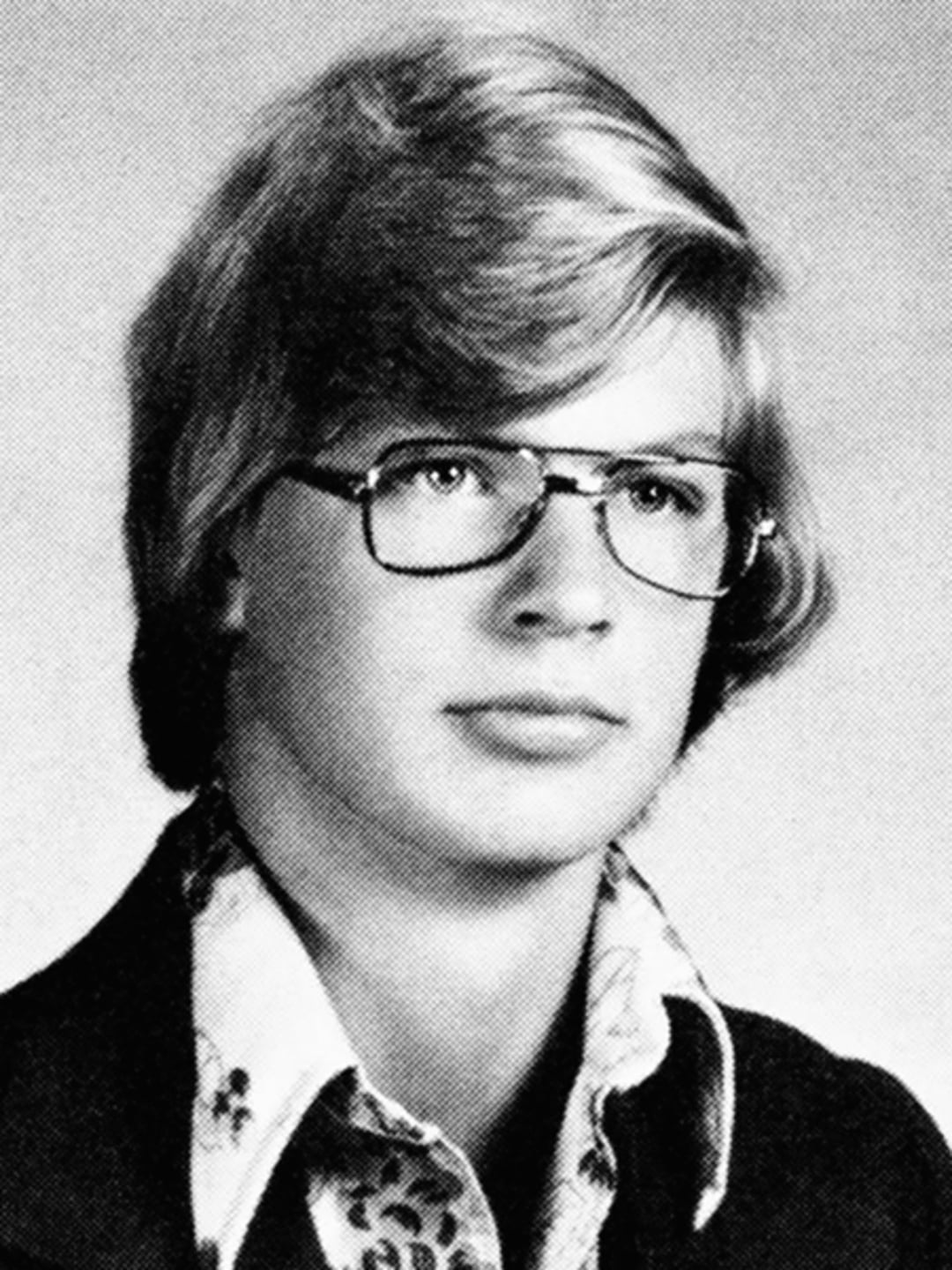 Black and white yearbook photo of a young person with light-colored, medium-length hair, wearing large eyeglasses, a patterned collared shirt, and a dark jacket, looking slightly to the side.