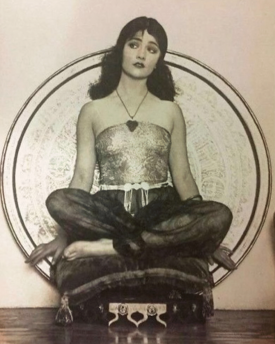 A woman in vintage attire sits cross-legged on a cushion against an ornate circular backdrop. She wears a strapless top, sheer pants, and a necklace, with her hair loose and an enigmatic expression on her face.