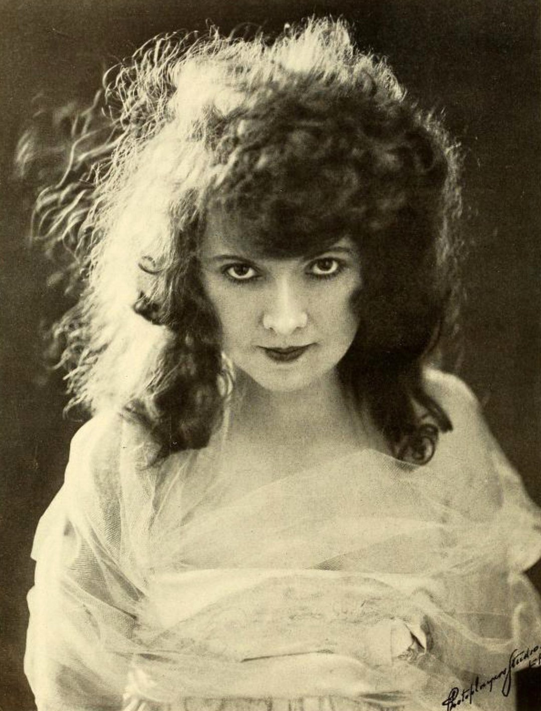 A woman with voluminous, curly hair and intense eyes looks directly at the camera. She wears a sheer, light-colored garment and stands against a dark background. The image is sepia-toned and softly lit.