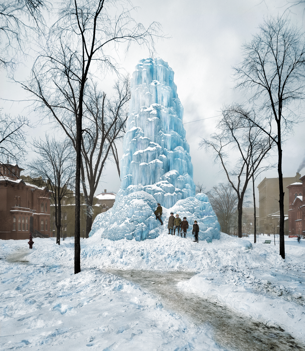 A tall, tower-like structure made of ice stands in a snowy park, surrounded by bare trees and several people gathered at its base. Nearby buildings are visible in the background under a cloudy sky.