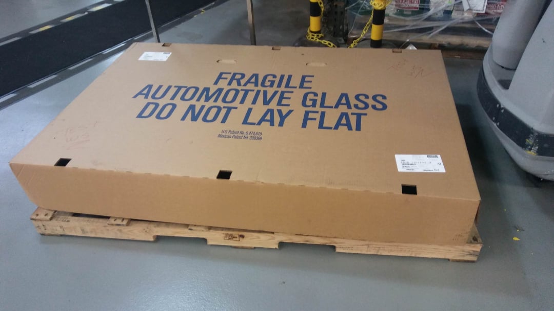 A large cardboard box labeled "FRAGILE AUTOMOTIVE GLASS DO NOT LAY FLAT" sits on a wooden pallet in an industrial setting, with machinery and yellow safety barriers in the background.