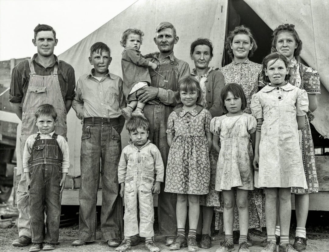 A black-and-white photo of a family, with adults and children of various ages, standing in front of a large canvas tent. The children wear simple, worn clothes and most look directly at the camera.