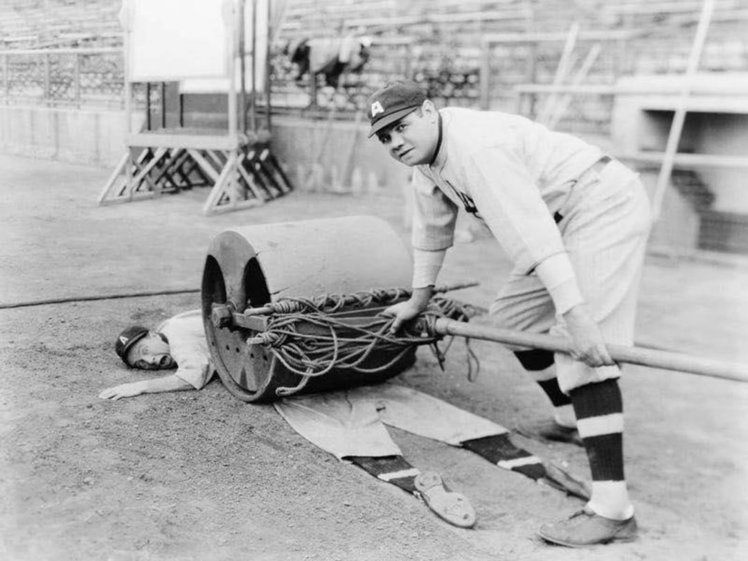 A baseball player in uniform looks at the camera while pushing a large field roller over another player lying flat on the ground, creating a humorous scene on an empty baseball field.
