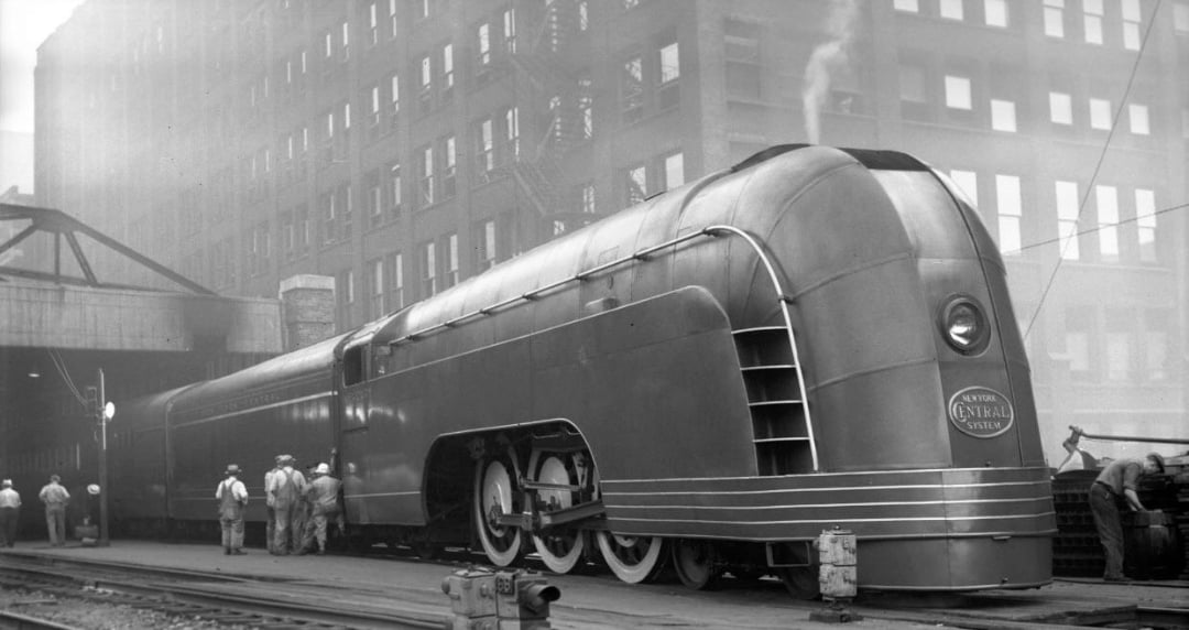 A vintage streamlined steam locomotive with a sleek, rounded design is parked at a station. Several people stand nearby amid tall, industrial buildings and light steam rising into the air.