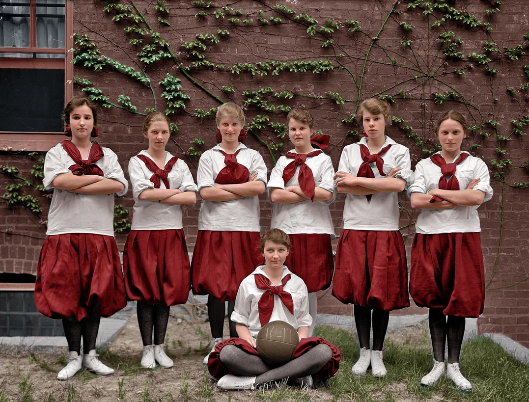 Seven young women in vintage gym uniforms with red skirts and white blouses pose in front of a brick wall covered in vines; six stand with arms crossed, while one sits cross-legged in front holding a volleyball.