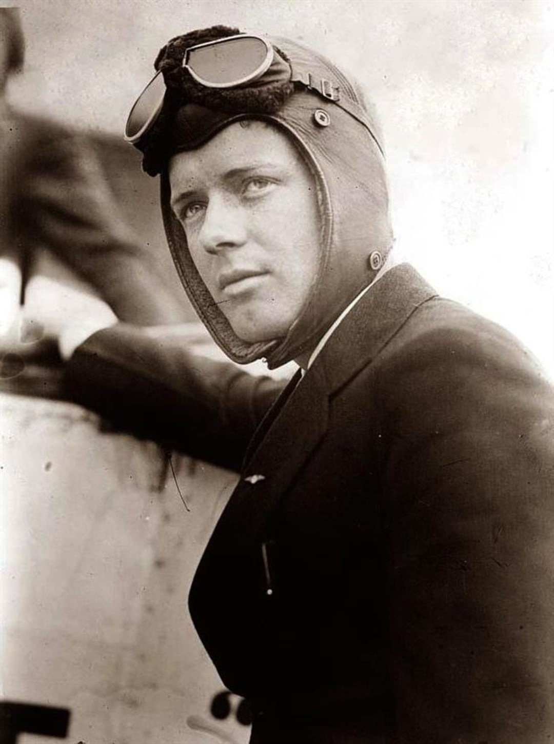 A black-and-white photo of a man wearing a vintage aviator cap and goggles, dressed in a suit, leaning on an aircraft and looking into the distance with a serious expression.