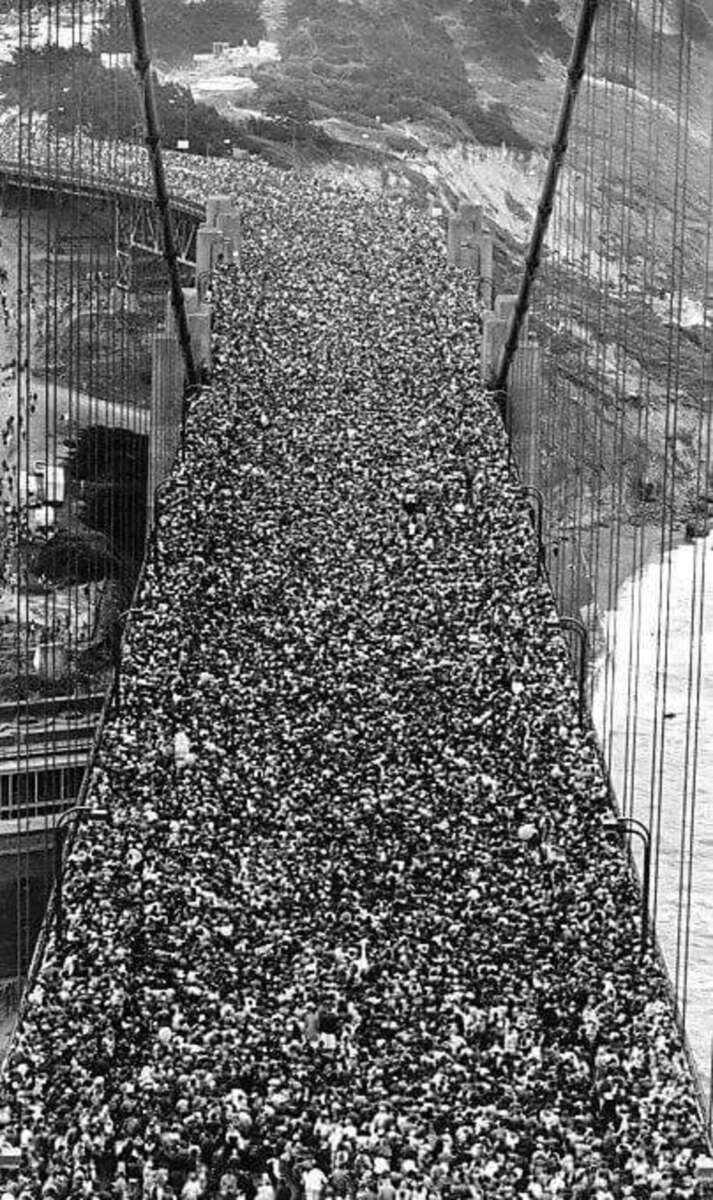 A massive crowd of people tightly packed together completely fills a suspension bridge, stretching from one end to the other, with rails and cables visible on the sides. The image is in black and white.