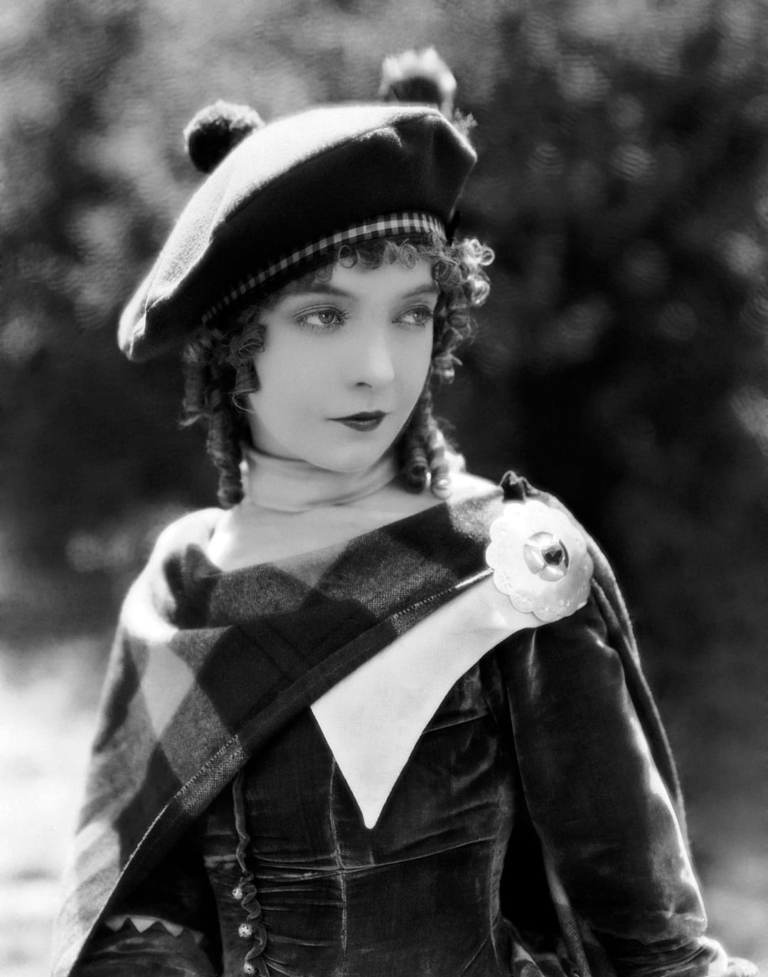 A woman in vintage clothing, wearing a checkered sash, a dark velvet dress, and a beret with two pom-poms, poses outdoors with a serious expression and curly hair. The photo is black and white.