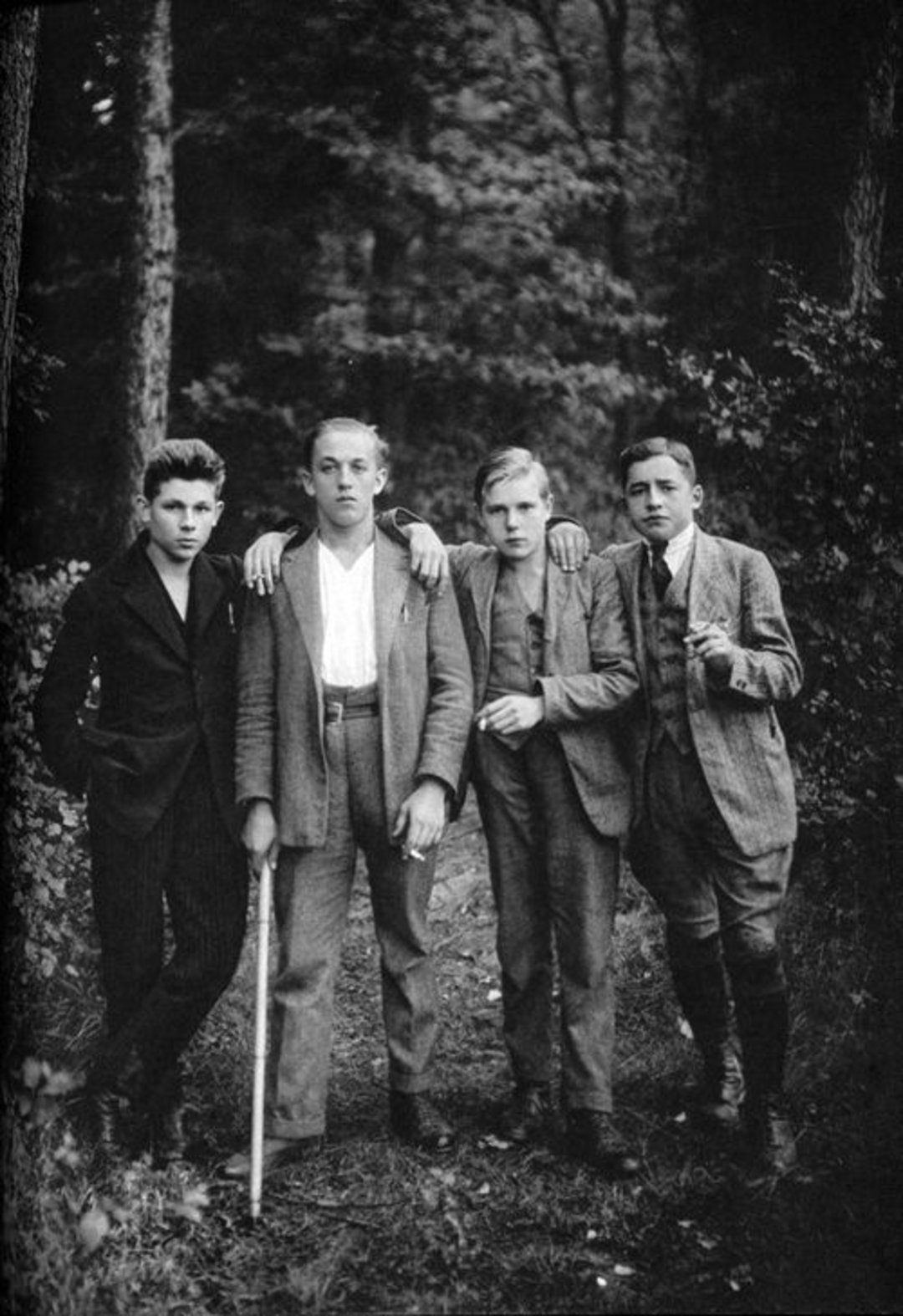 Four young men in old-fashioned suits stand together in a forest clearing. One has his arm around another and holds a cane. They look directly at the camera with serious expressions. The background is filled with trees and foliage.
