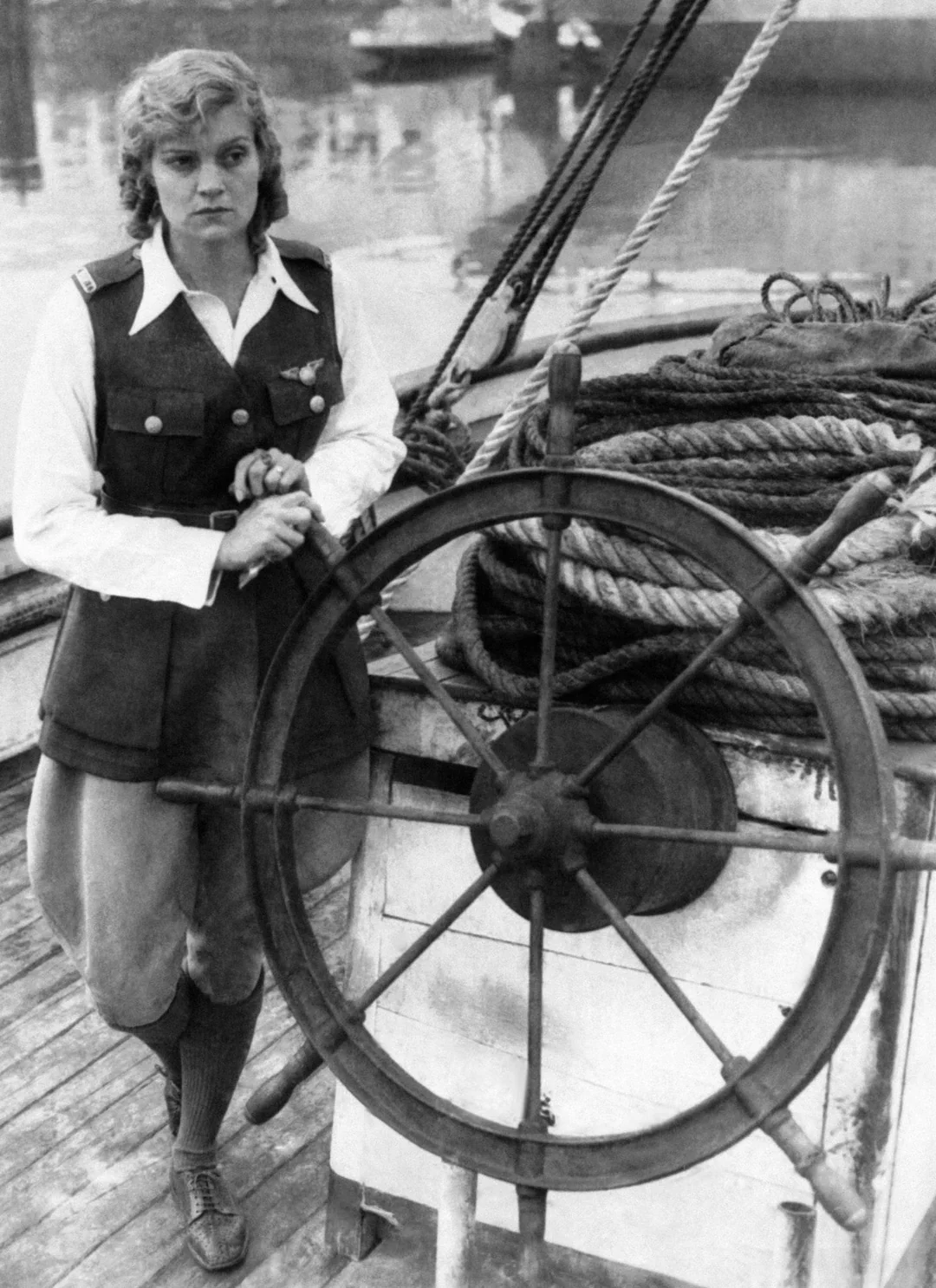 A woman in nautical attire stands by a ship’s wheel on a wooden deck, with coiled ropes and water in the background, looking directly at the camera.