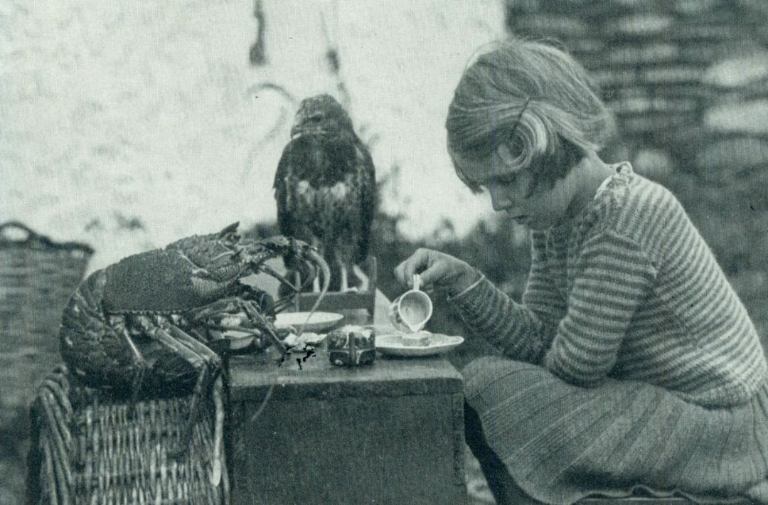 A young girl in a striped dress sits at a table, pouring tea into a cup. A large lobster and a bird perch on a basket nearby, appearing to join her for tea. The scene is in black and white.