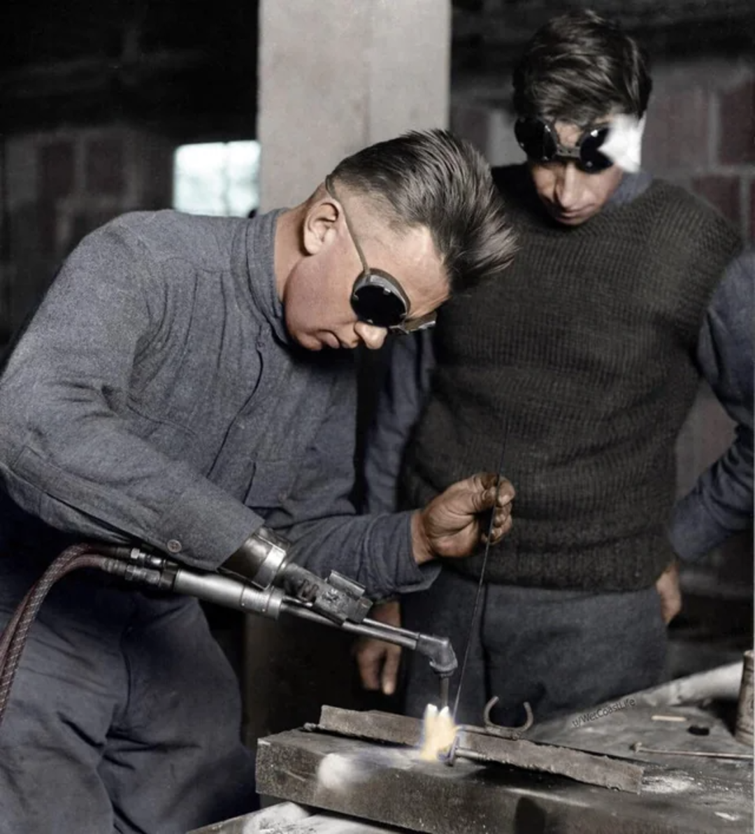 Two men work with a welding torch; one welds while wearing dark protective goggles, and the other stands beside him, also wearing eye protection. Both appear focused on the metalwork in front of them.