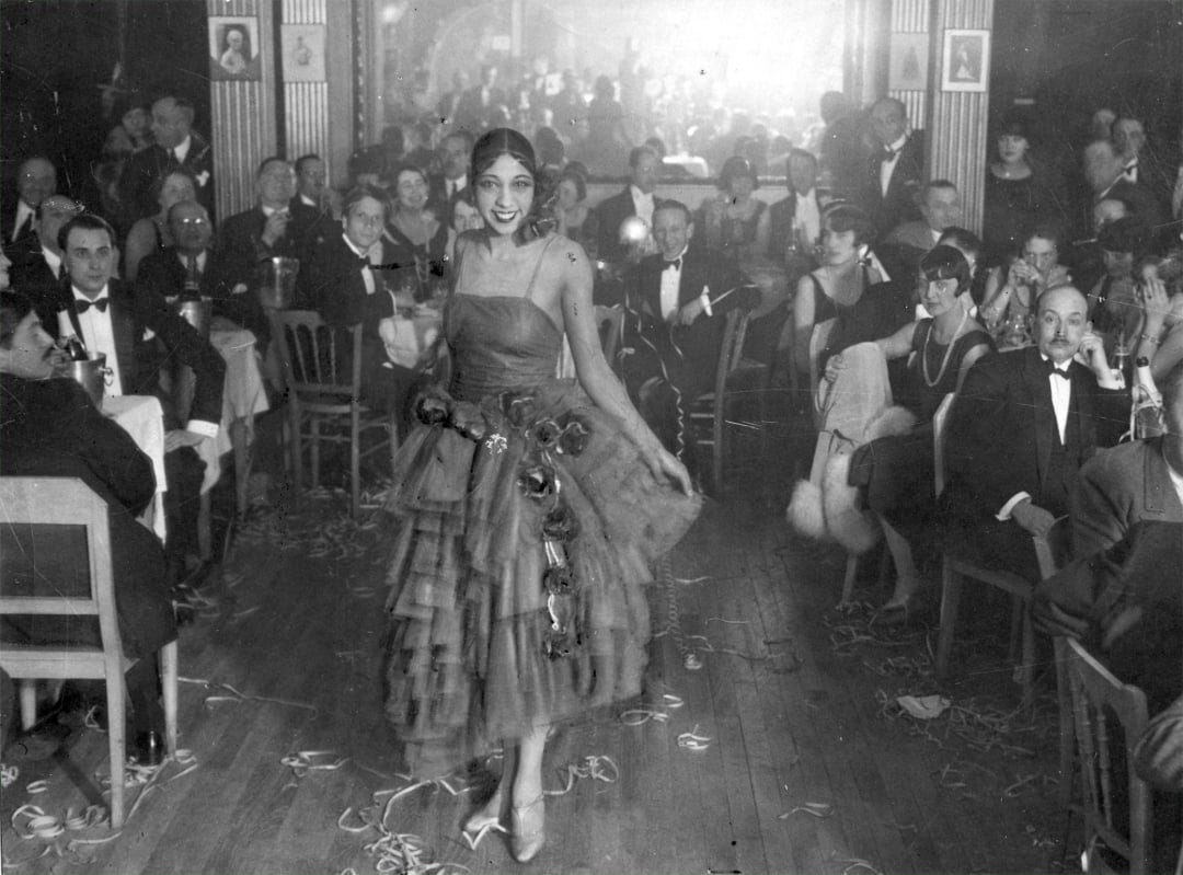 A woman in a ruffled dress performs on a dance floor before a large, seated audience in formal attire at a lively, crowded nightclub or cabaret in the early 20th century.