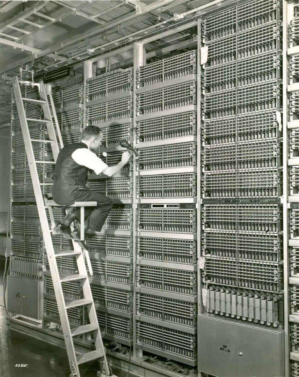 A man in a vest sits on a ladder adjusting wiring on a large wall of complex electrical equipment and relays, likely part of an early computer or telephone system.