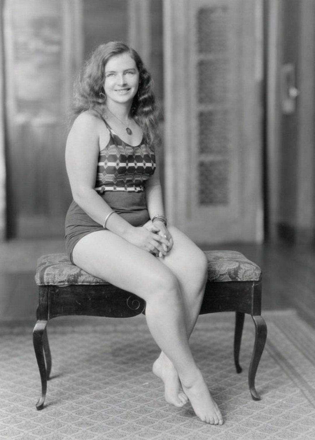 A young woman with wavy hair sits barefoot on a cushioned bench, wearing a patterned one-piece swimsuit and smiling at the camera. The background shows wooden doors and a patterned rug. The photo is in black and white.