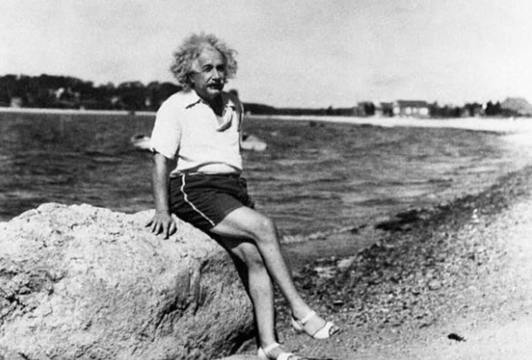 A man with wild hair sits on a large rock by a beach, wearing a light shirt, dark shorts, and sandals. The shoreline and distant buildings are visible in the background under a bright sky.