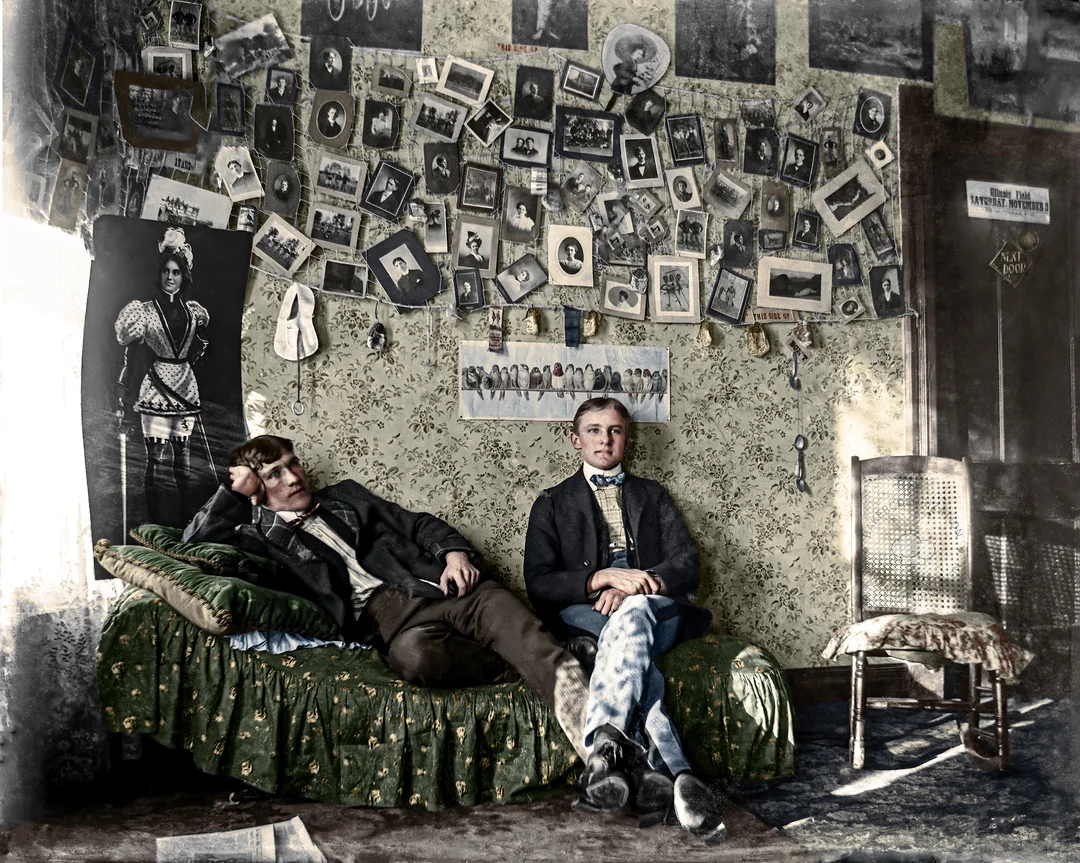 Two young men sit in a vintage room with patterned wallpaper, one lounging on a green couch and the other sitting upright. The wall behind them is covered with numerous old photographs and portraits. A wooden chair sits nearby.